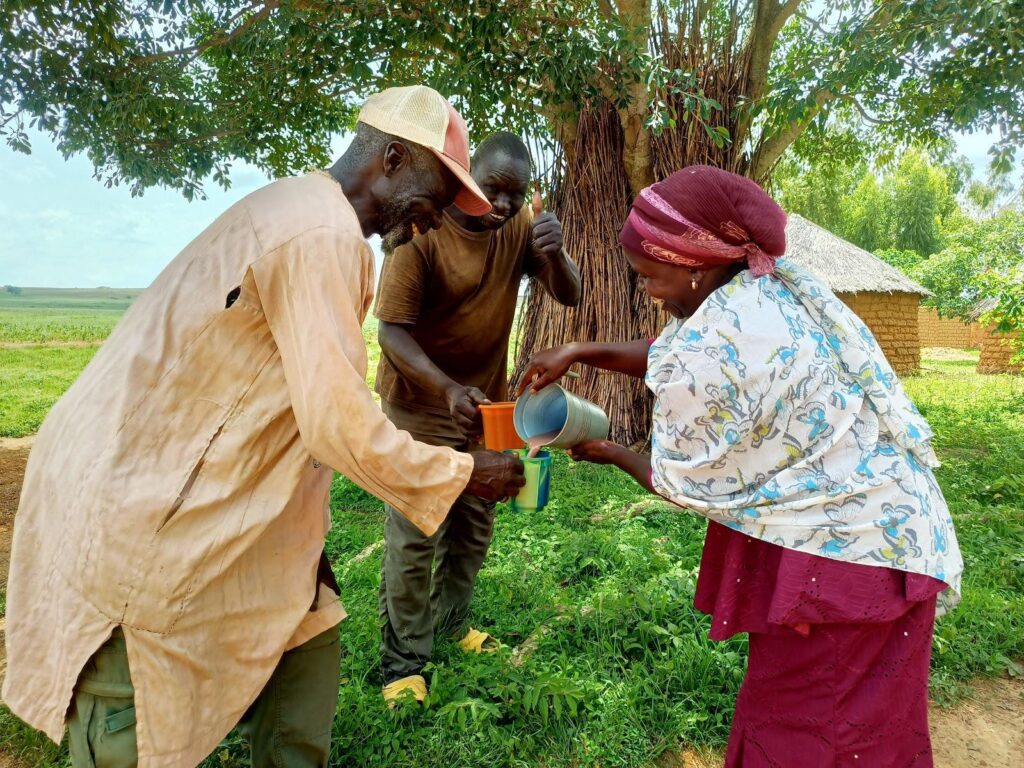 Three adults sharing a drink under a tree in a rural setting.