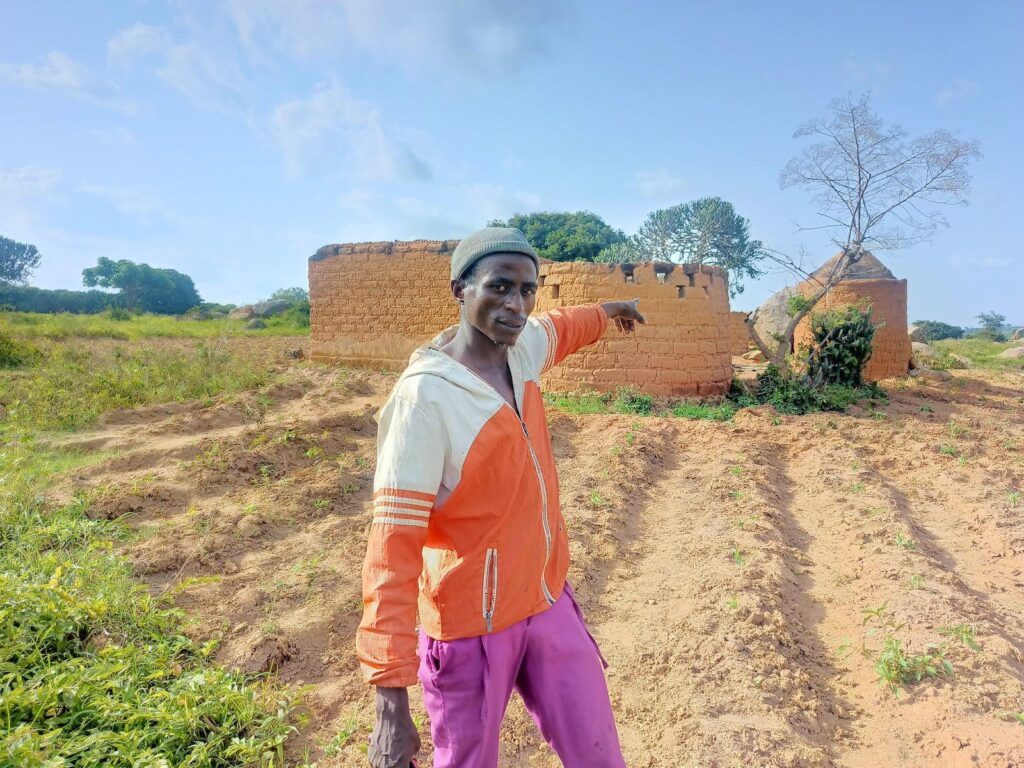 A man in a high-visibility shirt pointing at traditional mud brick structures under a blue sky.