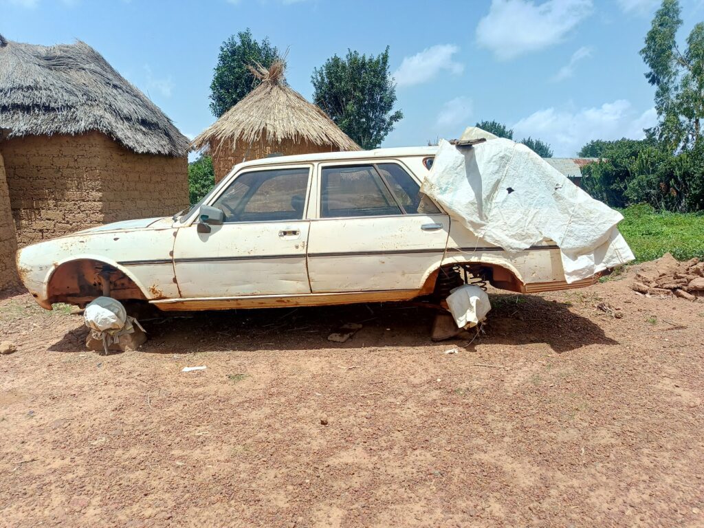 An old, abandoned car with missing wheels covered with a tarp, parked near traditional thatched-roof huts.