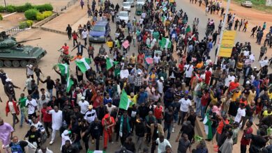 A large crowd of demonstrators with flags and banners marches on a street beside a military tank.