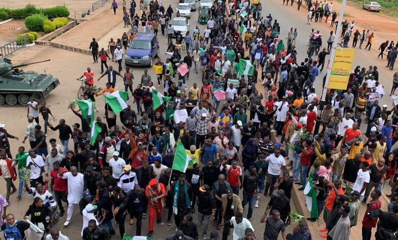 A large crowd of demonstrators with flags and banners marches on a street beside a military tank.