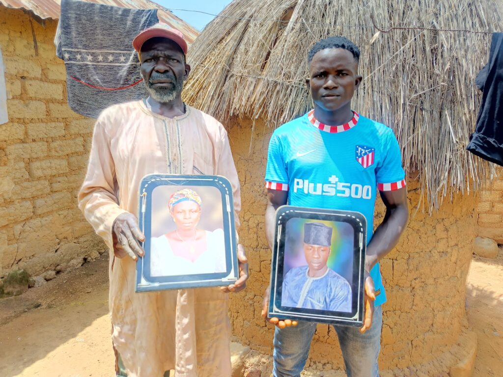 Two men standing outside a thatched hut, each holding a framed portrait.