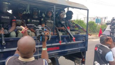 Armed police officers in a truck, being filmed by members of the press.