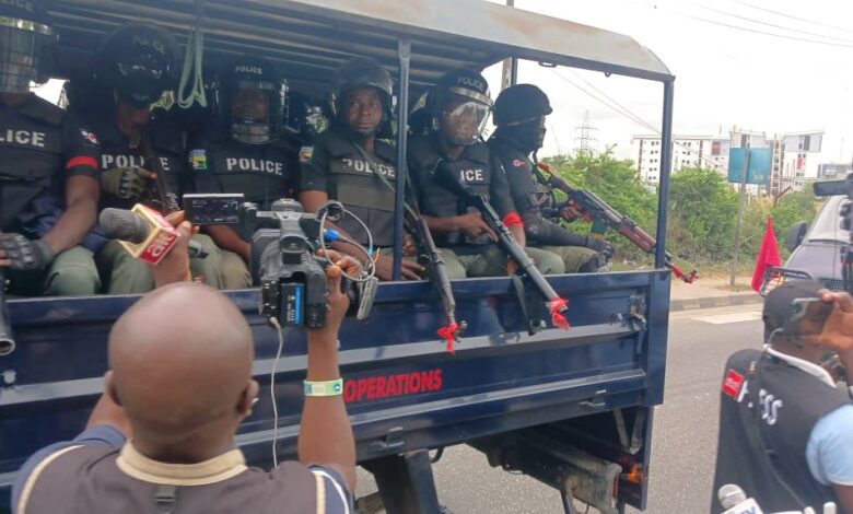 Armed police officers in a truck, being filmed by members of the press.