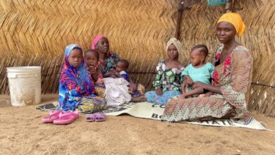 A group of women and children sitting together in front of a thatched wall, with colorful traditional clothing.