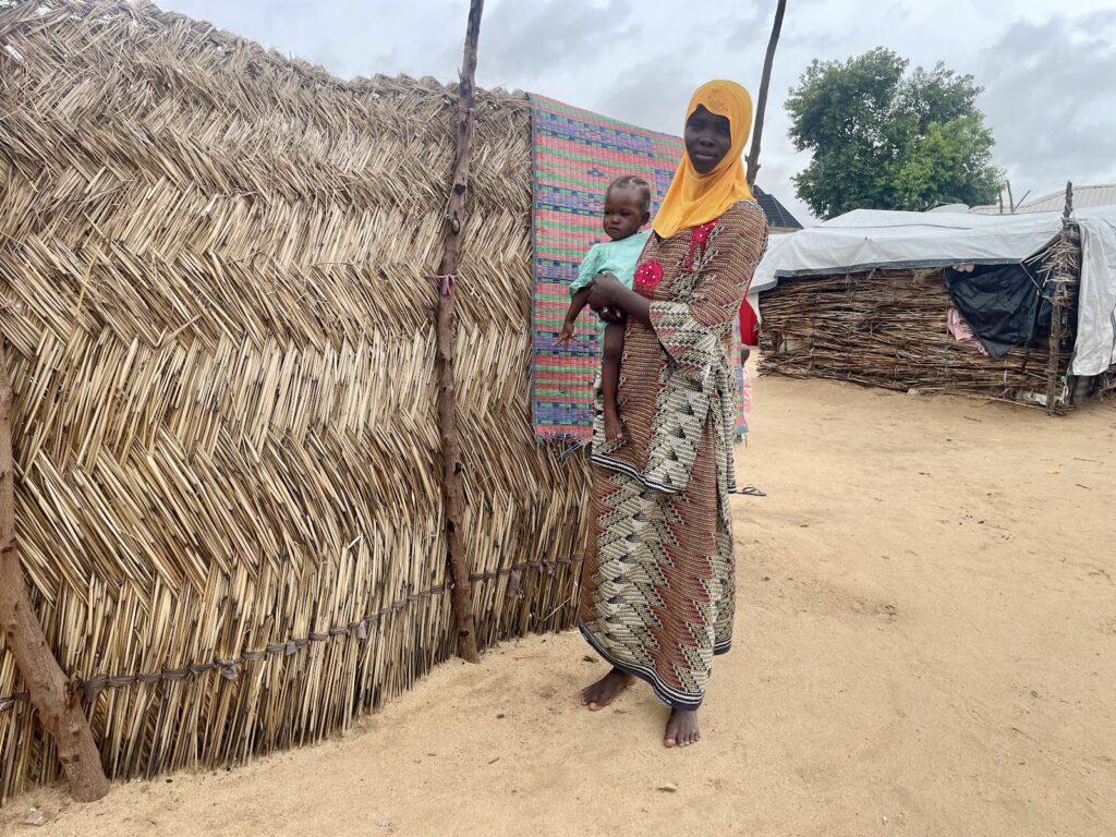 Woman in traditional attire holding a child, standing beside a thatched structure.