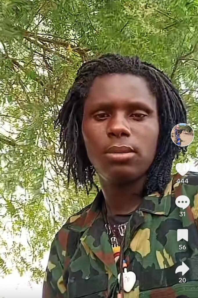 A person with braided hair and camouflage attire standing in front of green foliage.