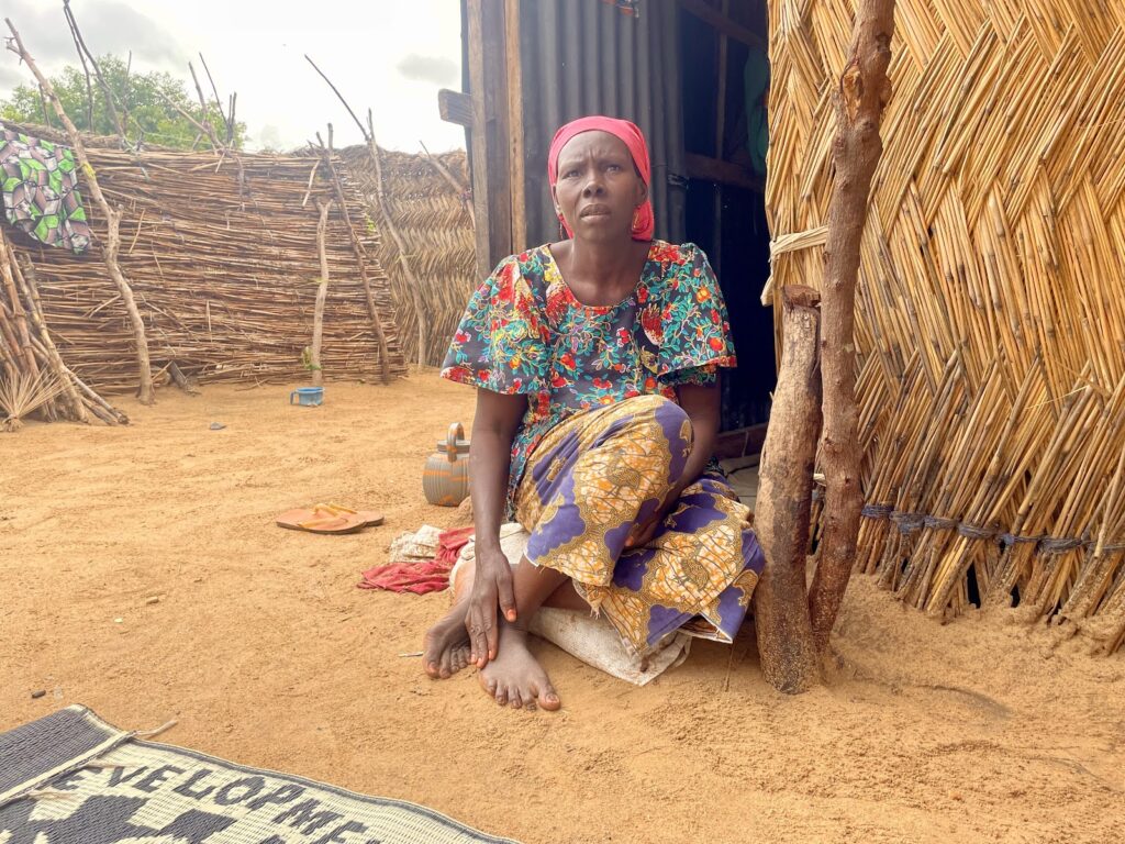 Woman sitting by a hut with thatched walls, looking thoughtful, in a rural setting.