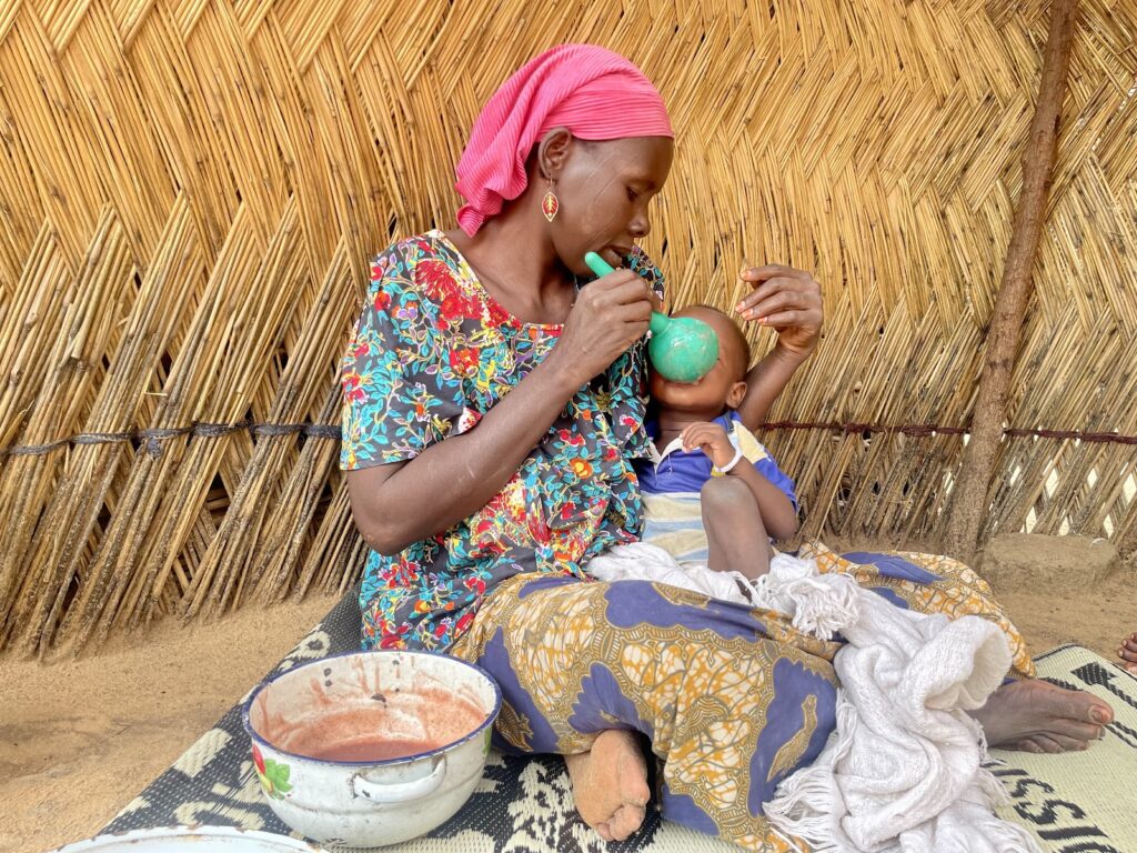 Woman in colorful clothing feeding a child with a spoon, sitting on a mat inside a hut with a thatched wall.