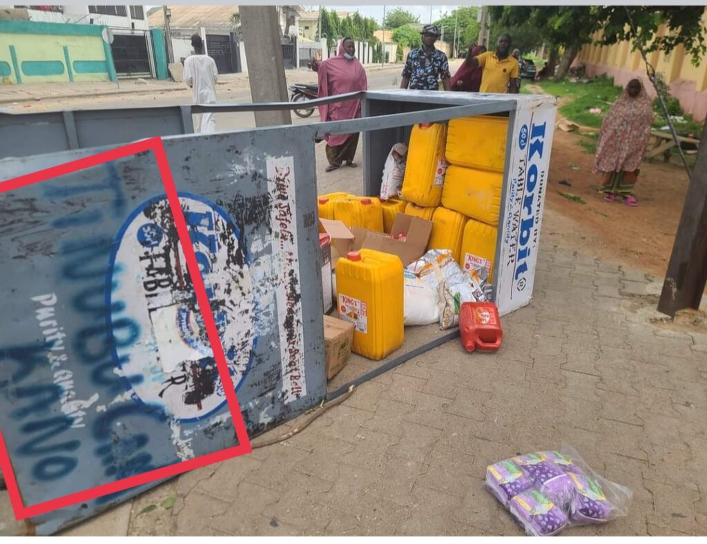 Overturned street vendor's kiosk with products scattered outside and onlookers in the background.