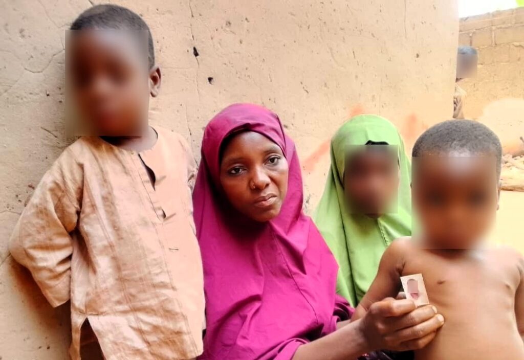 Woman in a pink headscarf sitting between two blurred face children, with another in front holding a bandage.