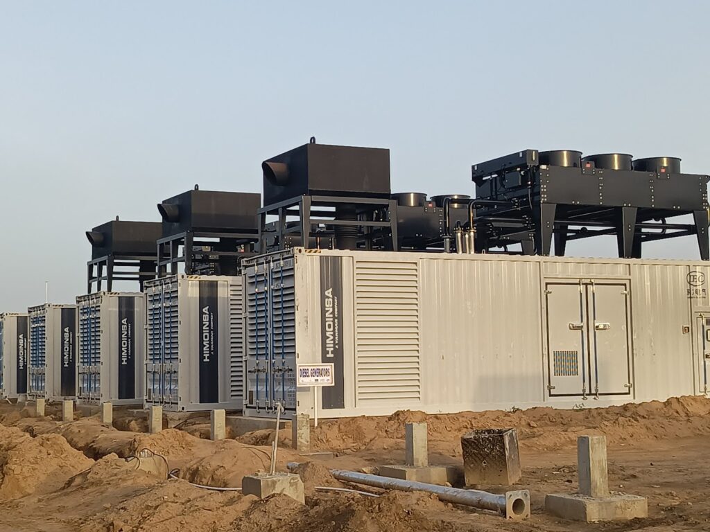 Industrial site with multiple large batteries and cooling equipment on a sandy ground under a clear sky.