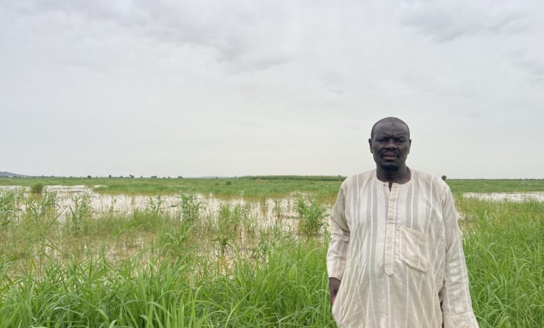 Man in traditional attire standing before a green field with a cloudy sky.