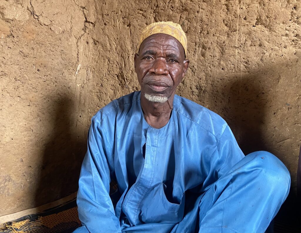 Elderly man in blue attire sitting against a mud wall.