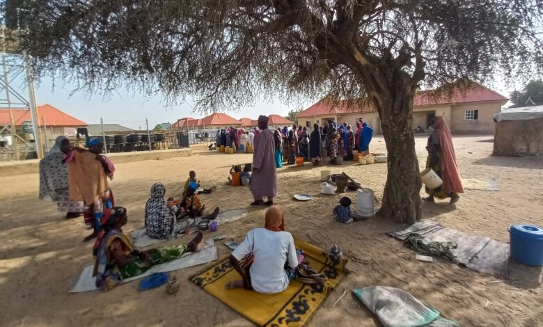 People gathering under a tree in a sandy area with mats, containers, and buildings in the background.