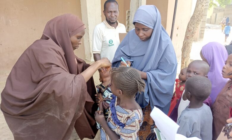 Health workers in hijabs administering care to children in an outdoor setting with onlookers.