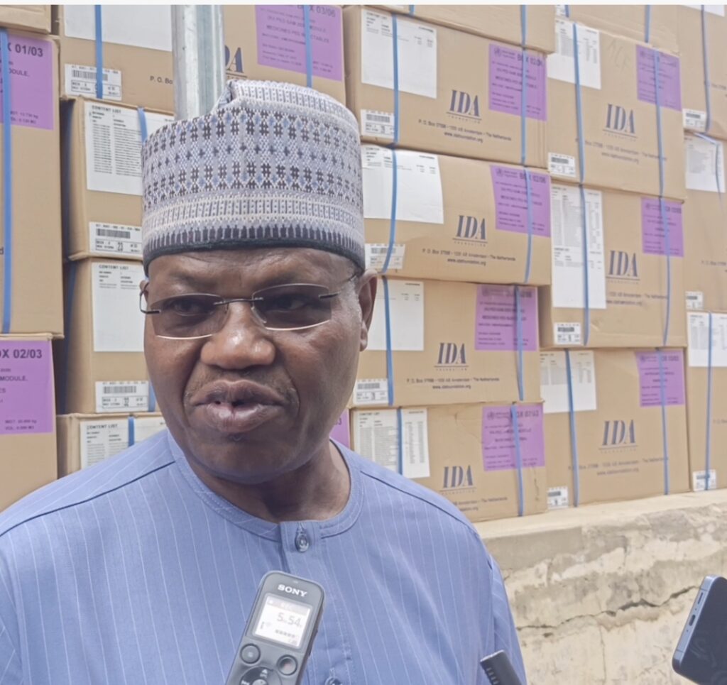 Man in traditional cap speaking into microphone with stacked boxes in the background.