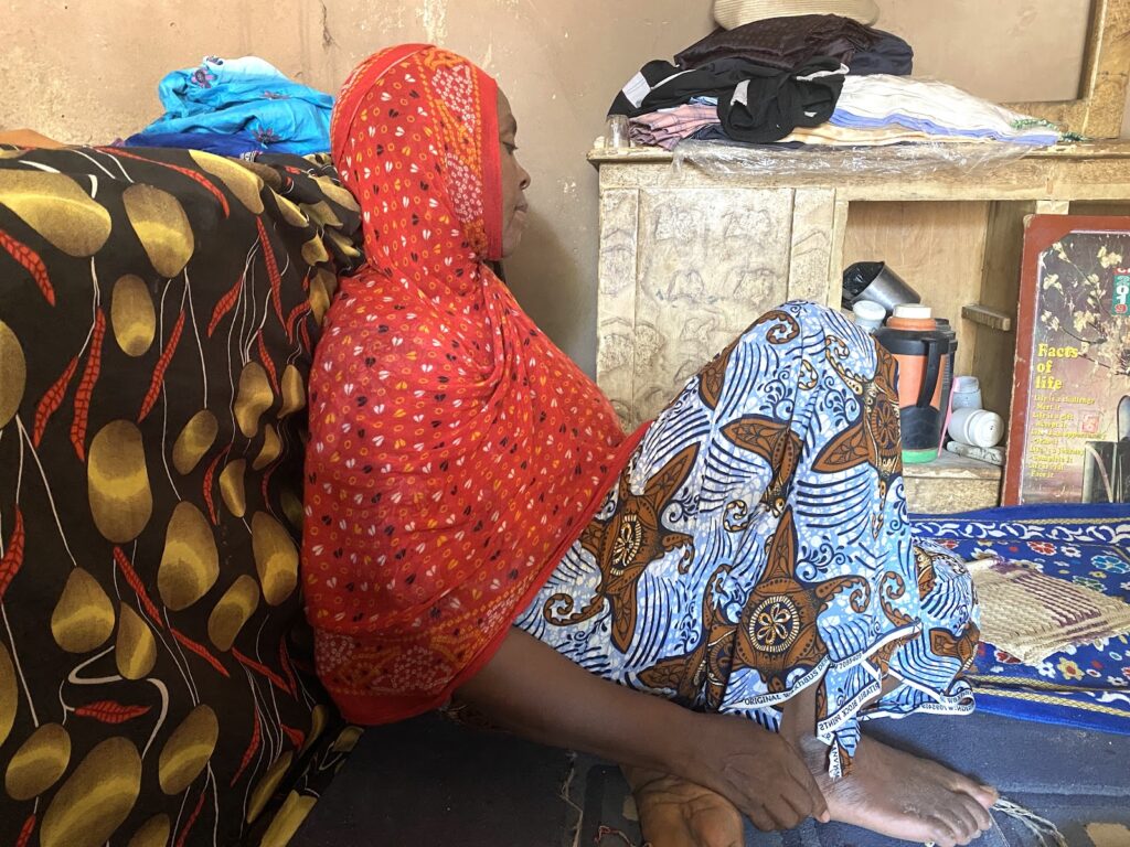 Two people in colorful traditional attire sitting in a room with clothes and personal items around.