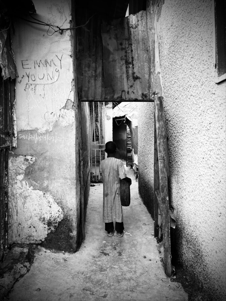 A person standing in a narrow alley with worn walls and graffiti, captured in black and white.