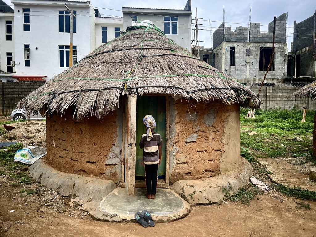 Person standing in the doorway of a traditional thatched-roof mud hut with modern buildings in the background.