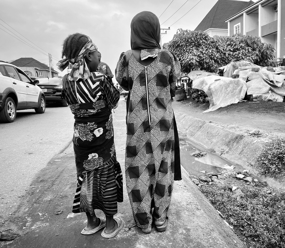 Two girls in traditional dresses standing by a roadside, with cars and houses in the background.