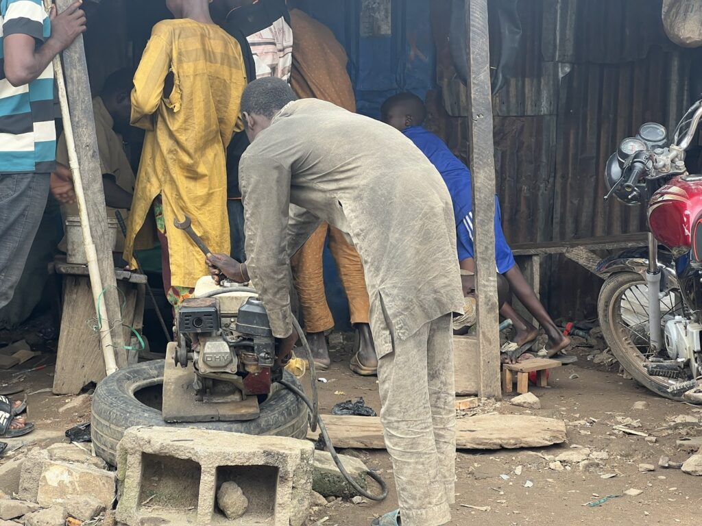 Man working on a generator at a busy outdoor workshop with people and motorcycles around.