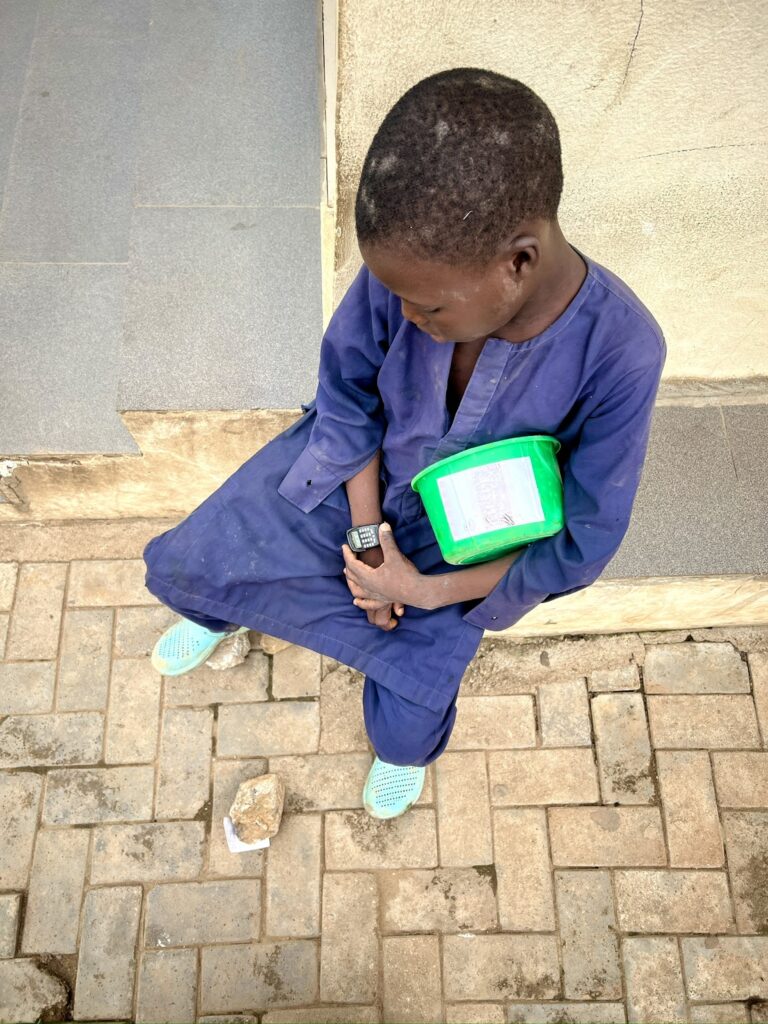 Young person sitting on a sidewalk, looking at a wristwatch, with a notebook and rock nearby.