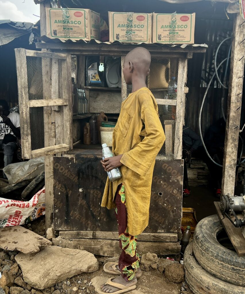 Young person in yellow robe holding bottles stands by a rustic wooden stall with various items.