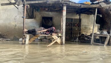 Person sleeping on makeshift bed above floodwaters near damaged house.