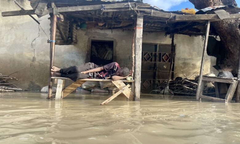 Person sleeping on makeshift bed above floodwaters near damaged house.