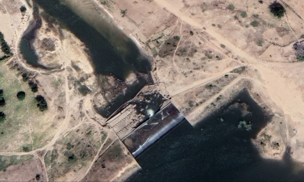 Aerial view of a dam with water flowing through its spillways into a river, surrounded by arid land.