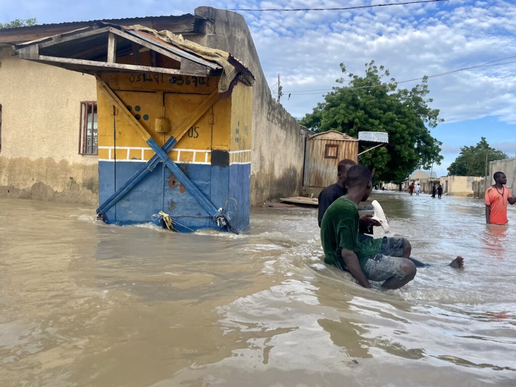 Flooded street with people wading through water near a damaged building with bracing supports.