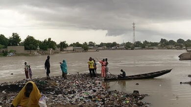 People beside a river with a boat on littered banks under cloudy skies.