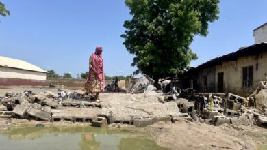 Woman walking among debris and stagnant water near dilapidated buildings under a clear sky.