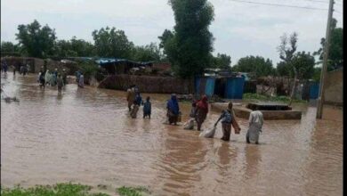 People wading through a flooded village street with houses partially submerged and possessions carried overhead.