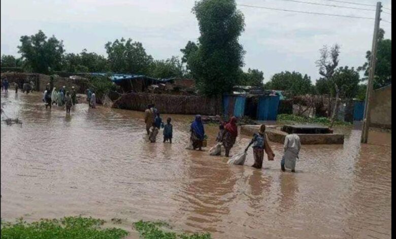 People wading through a flooded village street with houses partially submerged and possessions carried overhead.