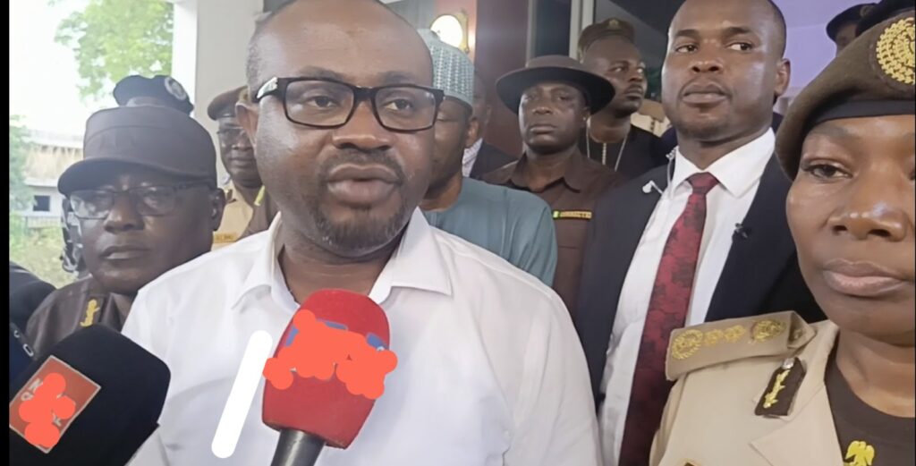 Man in glasses speaking into microphones with security personnel and officials in background.