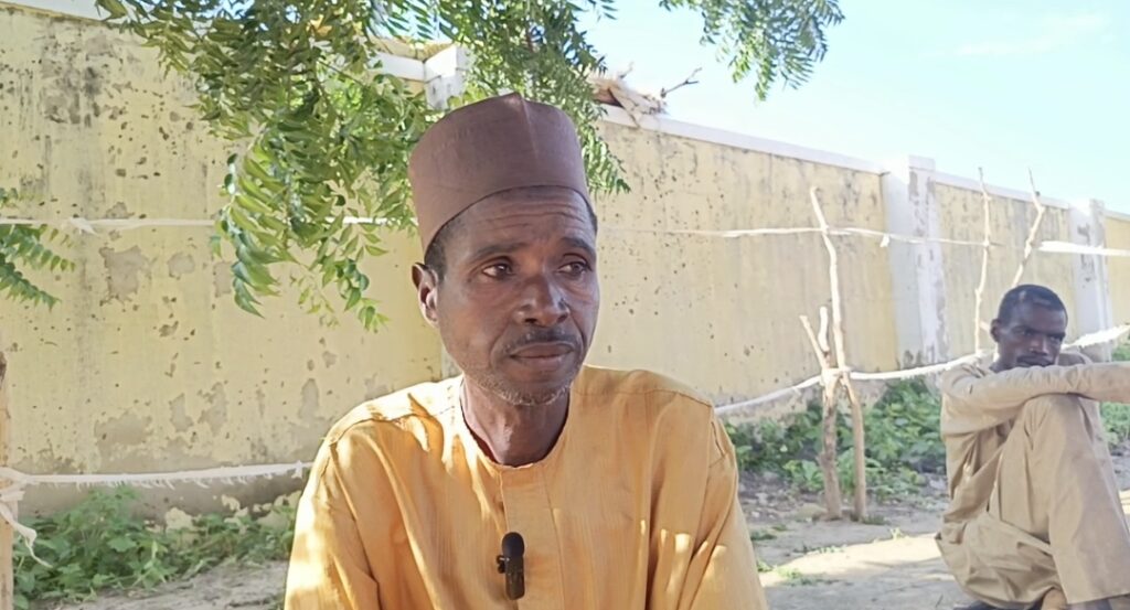 Man in yellow clothing with fez cap sitting outdoors, another person in background, against a wall with greenery.