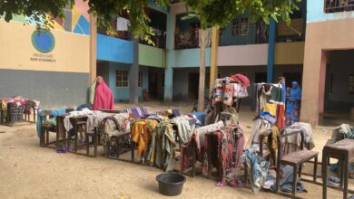 Colorful clothing laid out on tables and benches to dry in a sunlit courtyard, with women standing nearby.