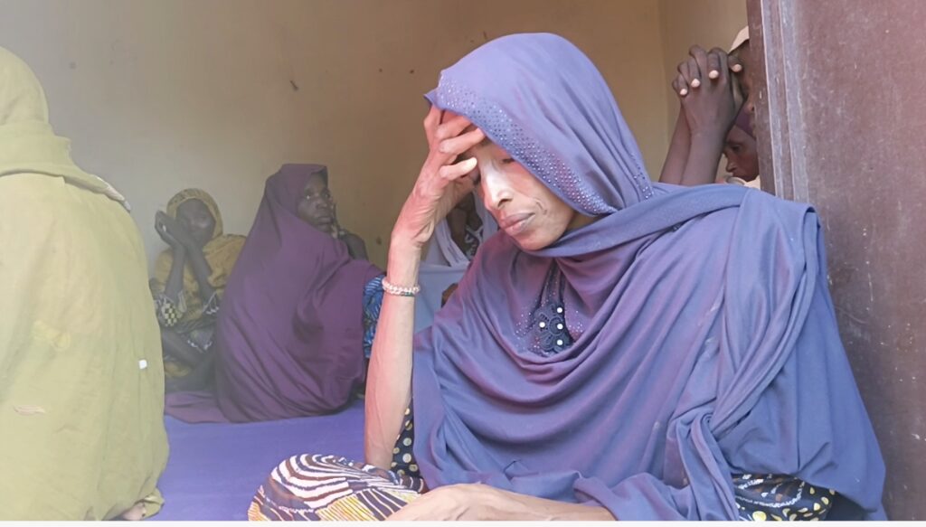 A person in a purple garment with a thoughtful expression, resting their forehead on their hand, indoors with others nearby.