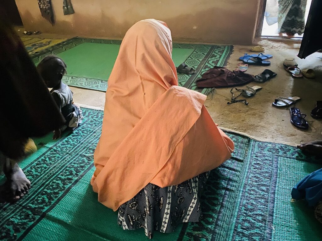 Person in orange hijab sitting on a green mat in a room with scattered sandals and a small child nearby.