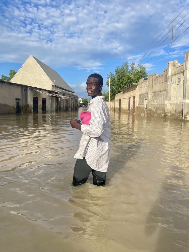 A person standing in a flooded street with buildings partially submerged in water, under a blue sky with clouds.