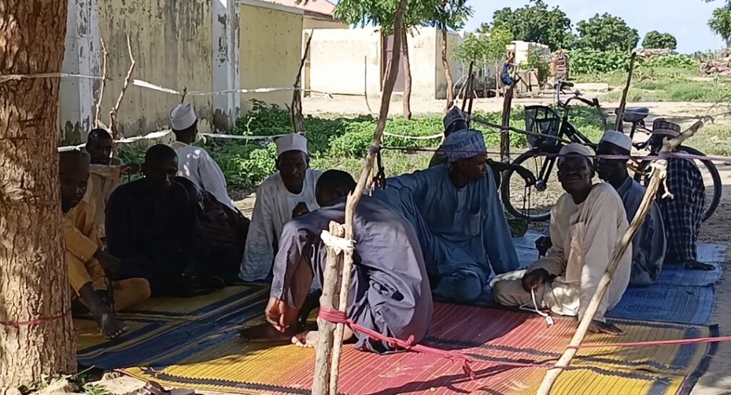 Men in traditional attire gather under a tree on woven mats, chatting with bicycles parked behind them.