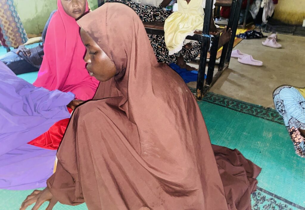 Woman in brown hijab sitting on floor with others in colorful clothing around her, indoors.