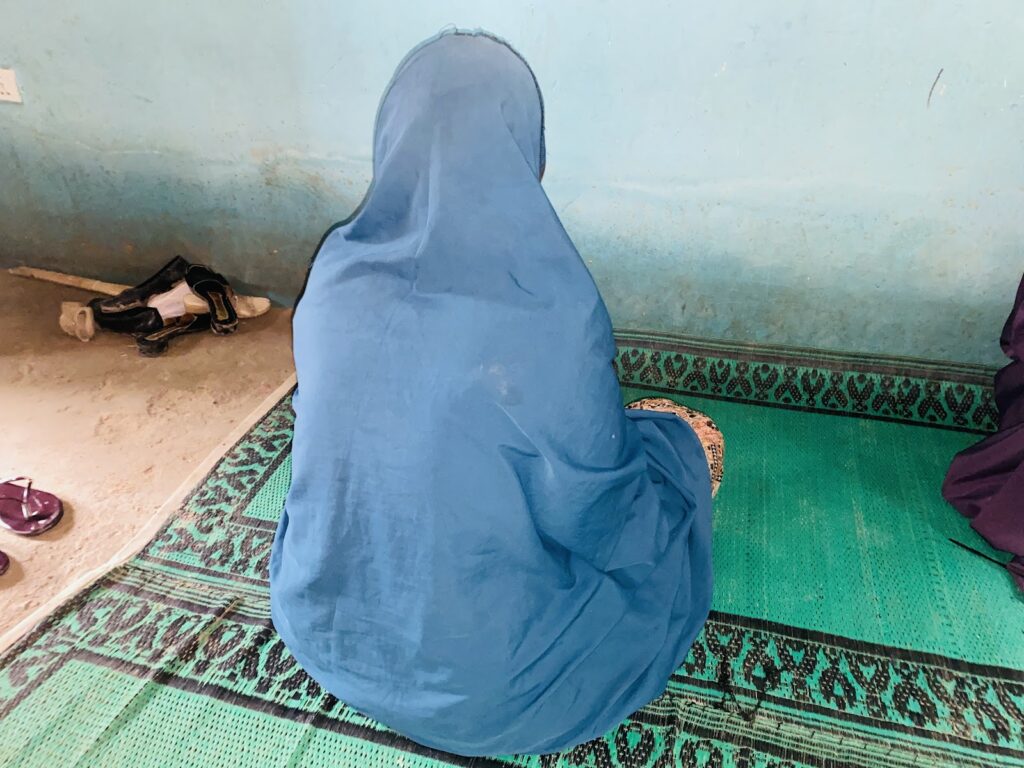 Person in blue attire seated on a green patterned rug against a faded blue wall, with shoes nearby.