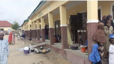 People outside a building with columns, some sitting, with pots and utensils around.