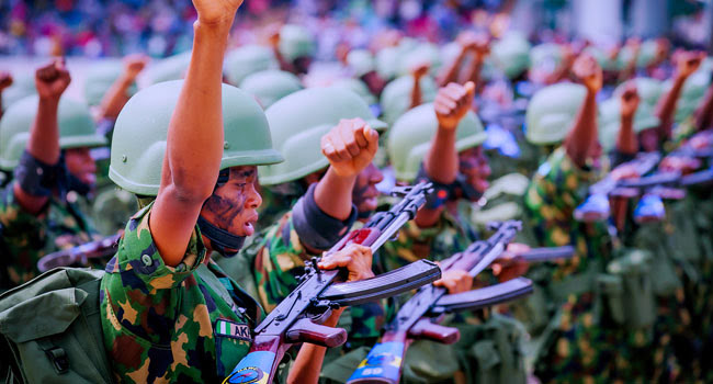 Uniformed soldiers with raised fists and rifles during a ceremony or rally.