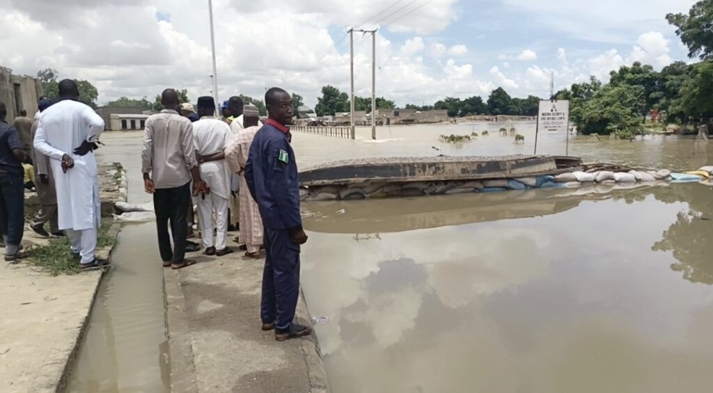 People observing a flooded area with a submerged bridge and sandbags, under a cloudy sky.