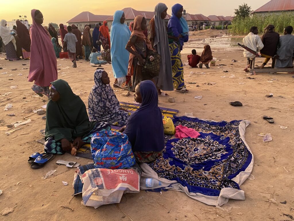 Women in traditional attire sitting on the ground in a crowded outdoor area at dust, with scattered litter.