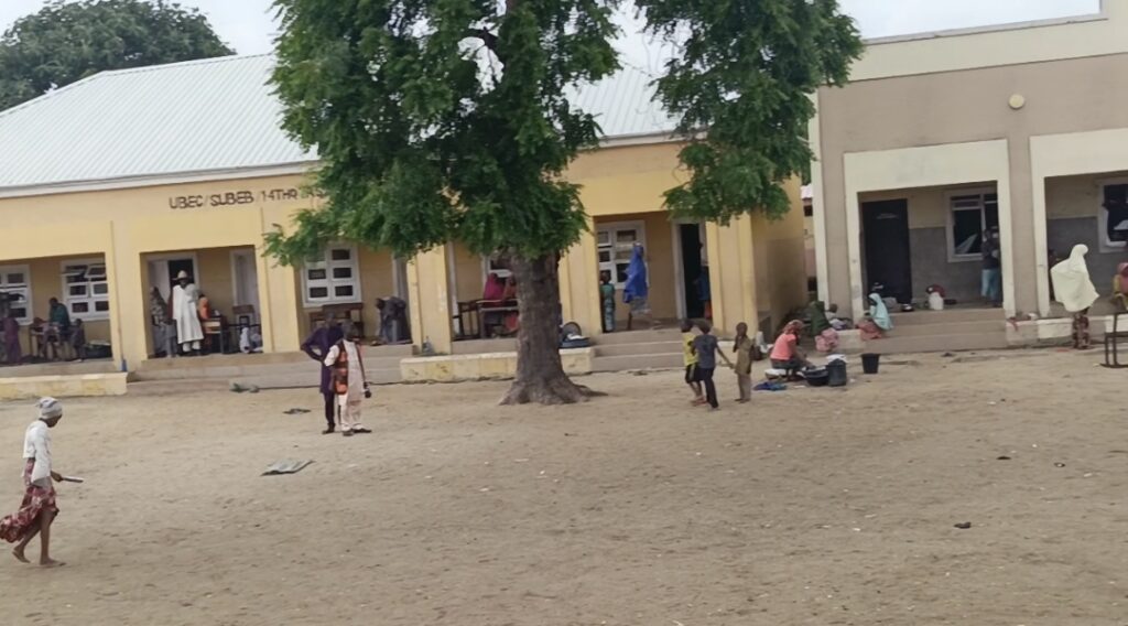 People outside a building with a sign "UBEC SUBEB 1T4R", under a tree in a sandy area.